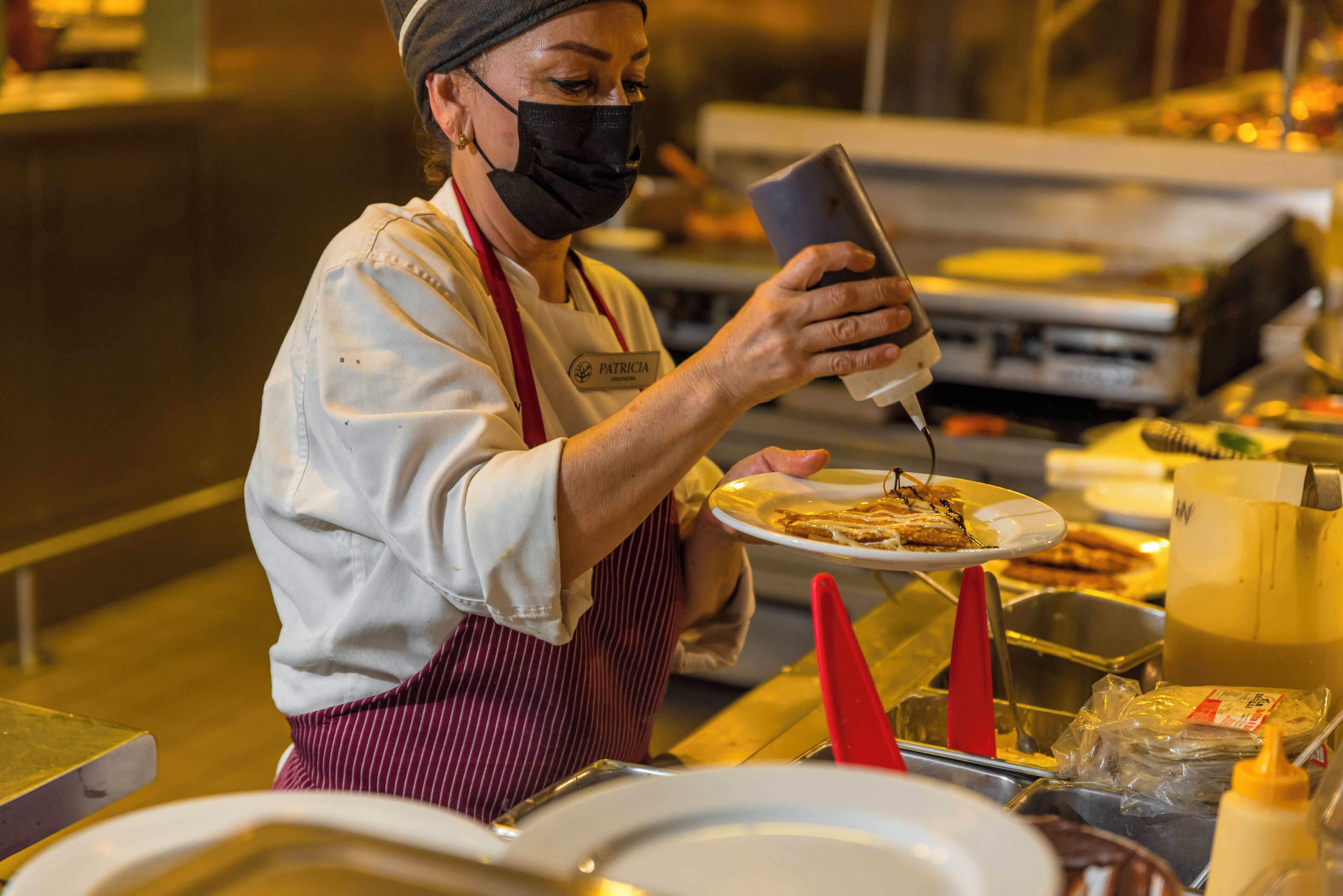 A person in a kitchen setting, wearing a white shirt and a red apron, holding a plate with food.