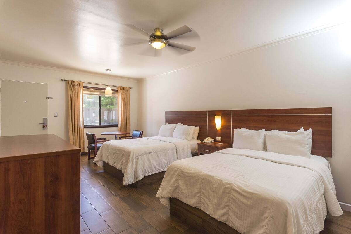 A hotel room with two beds, a desk, a chair, a wooden desk, a ceiling fan, and a window with curtains.