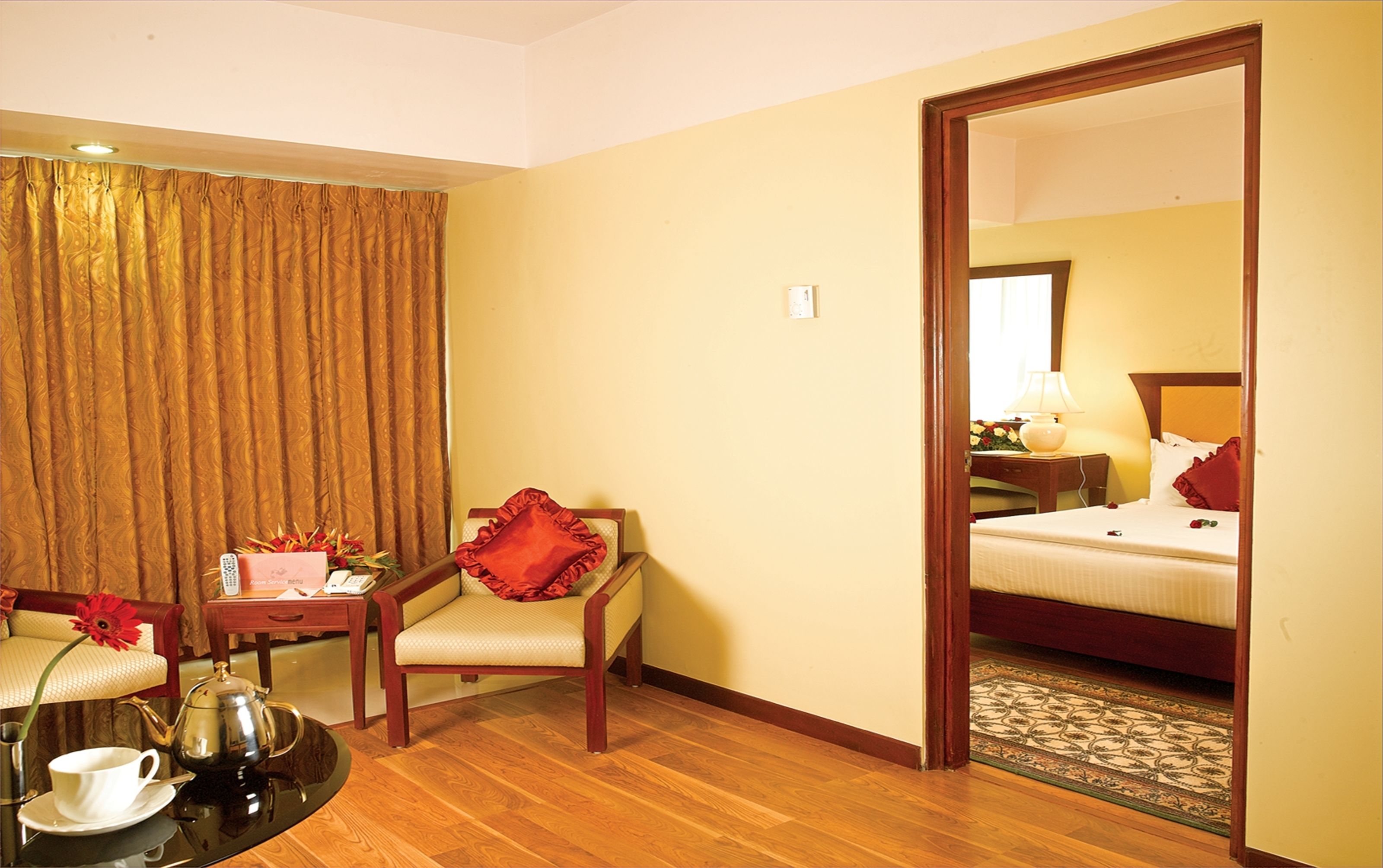 A hotel room with a wooden floor, a bed with white bedding, a red chair, a small table with a teapot and cups, and a mirror reflecting the room.