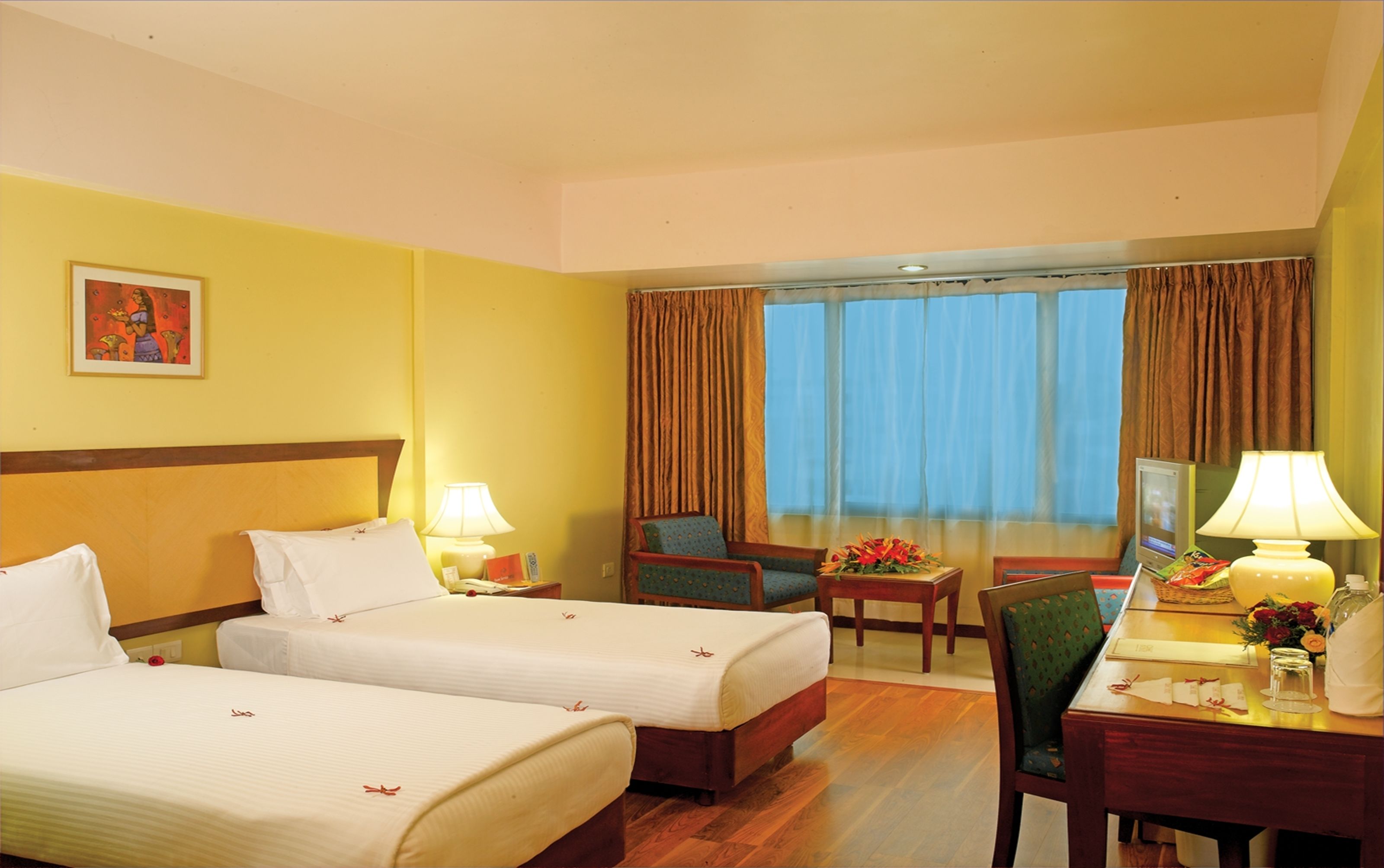 A hotel room with two beds, a desk with a lamp, a chair, a window with curtains, and a painting on the wall.