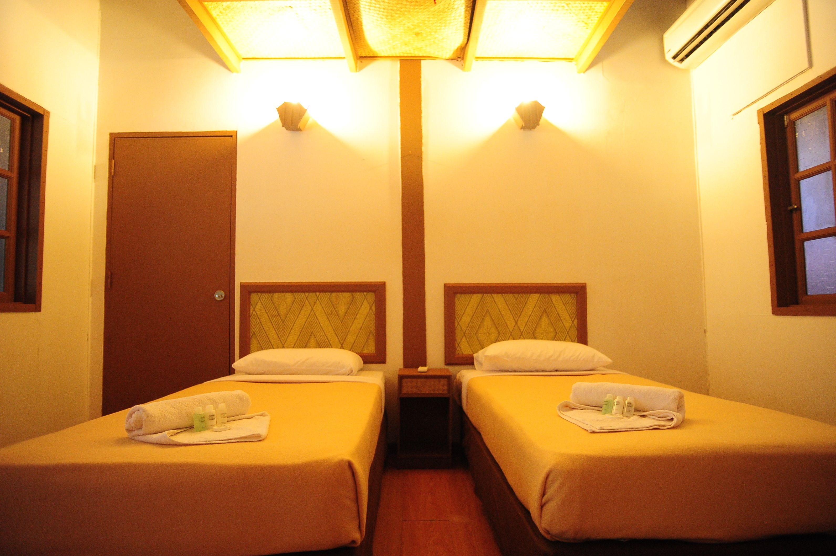 A hotel room with two single beds, a wooden door, and a small bedside table with a lamp.