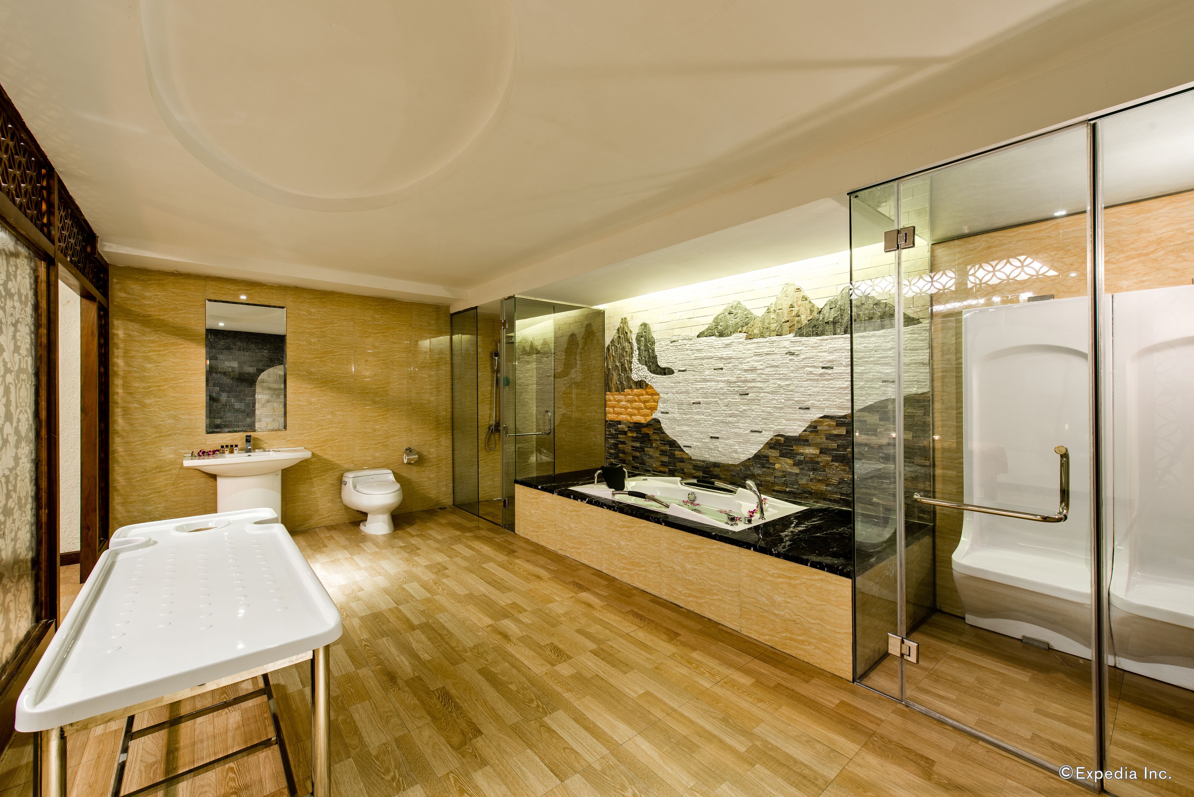 A modern bathroom with a wooden floor, a glass-enclosed shower, a white sink, and a bathtub with a stone facade.