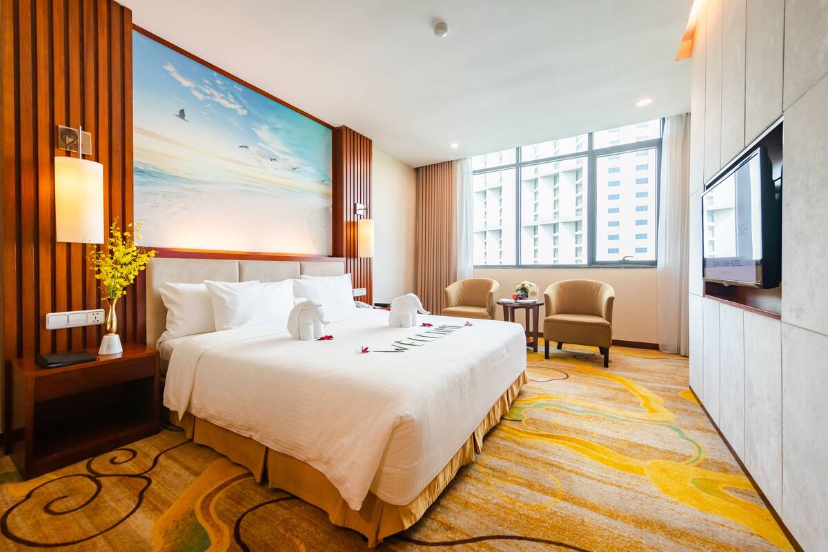 Ein Hotelzimmer mit einem großen Bett, einem Fernseher, einem Schreibtisch mit Lampe, einem Sessel und Blick auf die Stadt durch große Fenster.