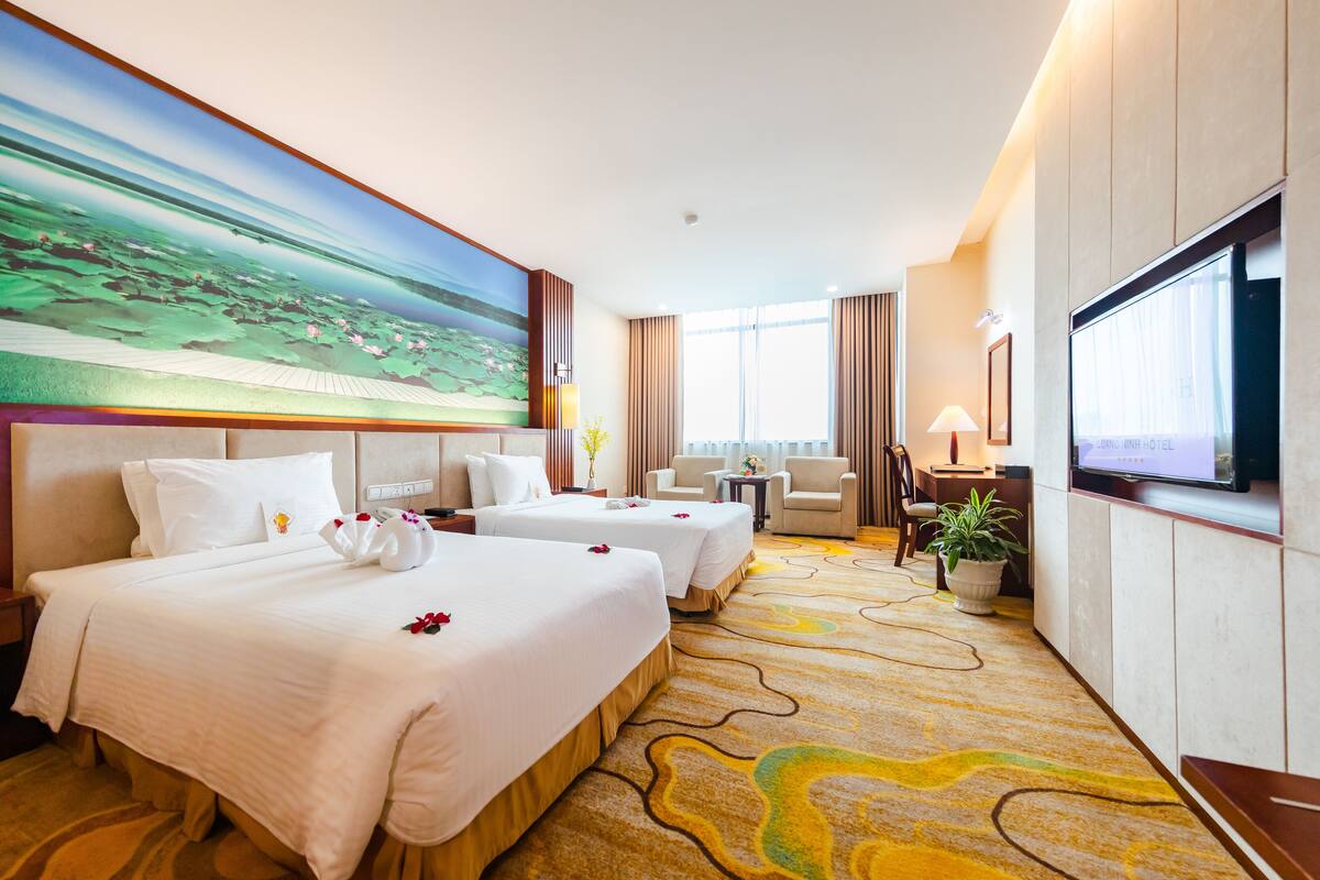 Ein Hotelzimmer mit zwei Betten, einem großen Gemälde, einem Fernseher, einem Schreibtisch, einem Sessel und einer Zimmerpflanze.