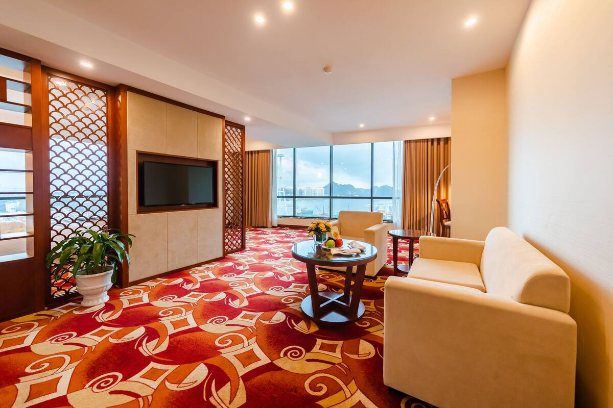 Ein modernes Hotelzimmer mit großem Fenster, einem Flachbildfernseher, einer Couch, einem kleinen runden Tisch mit einer Vase und einer Zimmerpflanze.