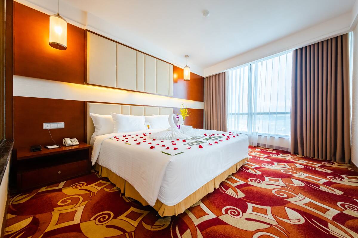 Ein Hotelzimmer mit einem großen Bett, einem Nachttisch mit Telefon, einem Fenster mit Vorhängen und einem gemusterten Teppich.