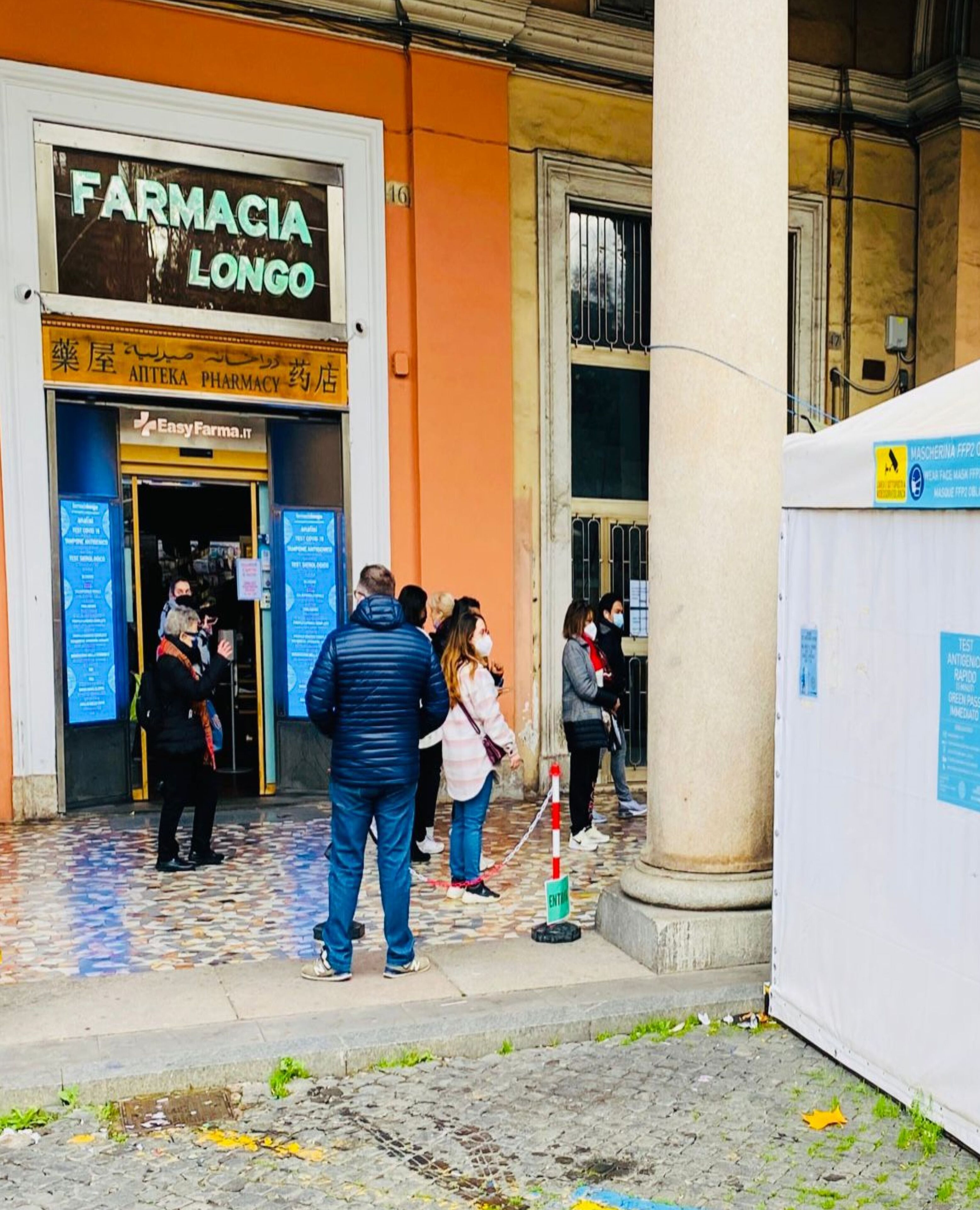 A pharmacy named 'Farmacia Longo' with a tiled entrance and people queuing outside.