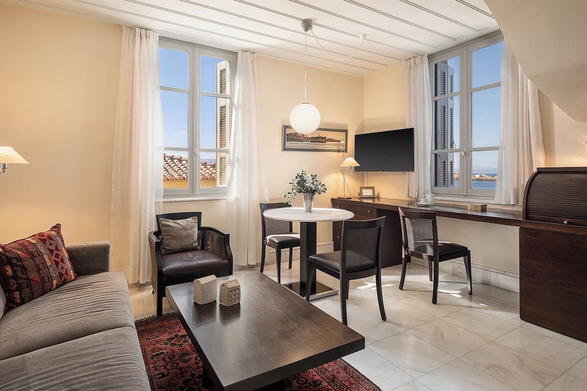 Una sala de estar moderna con sofá, mesa de centro y sillones. Cuenta con un comedor con mesa y sillas, un televisor montado en la pared y ventanas amplias con cortinas.