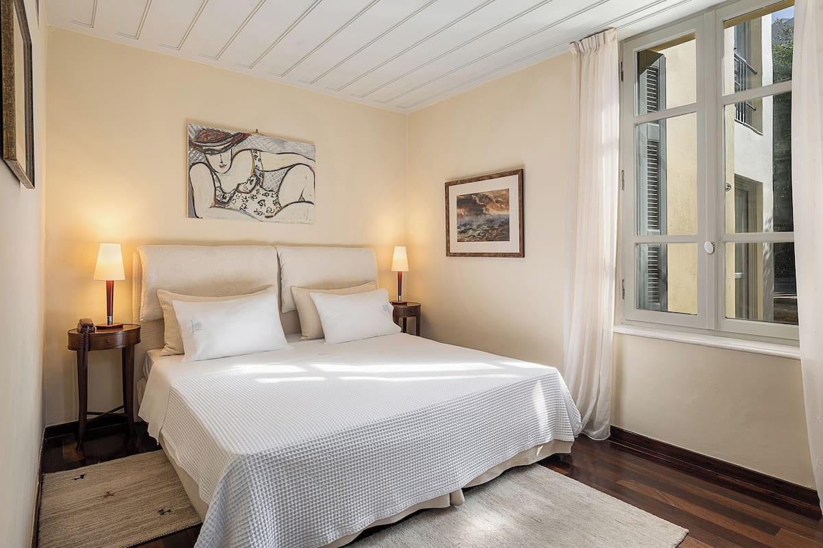 Un dormitorio ordenado con una cama, dos mesitas de noche, un buró, una ventana con contraventanas y obras de arte en las paredes.