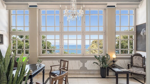 A room with large windows, a chandelier, a dining table, chairs, a sofa, and a potted plant. - Villa Andromeda (Chania)