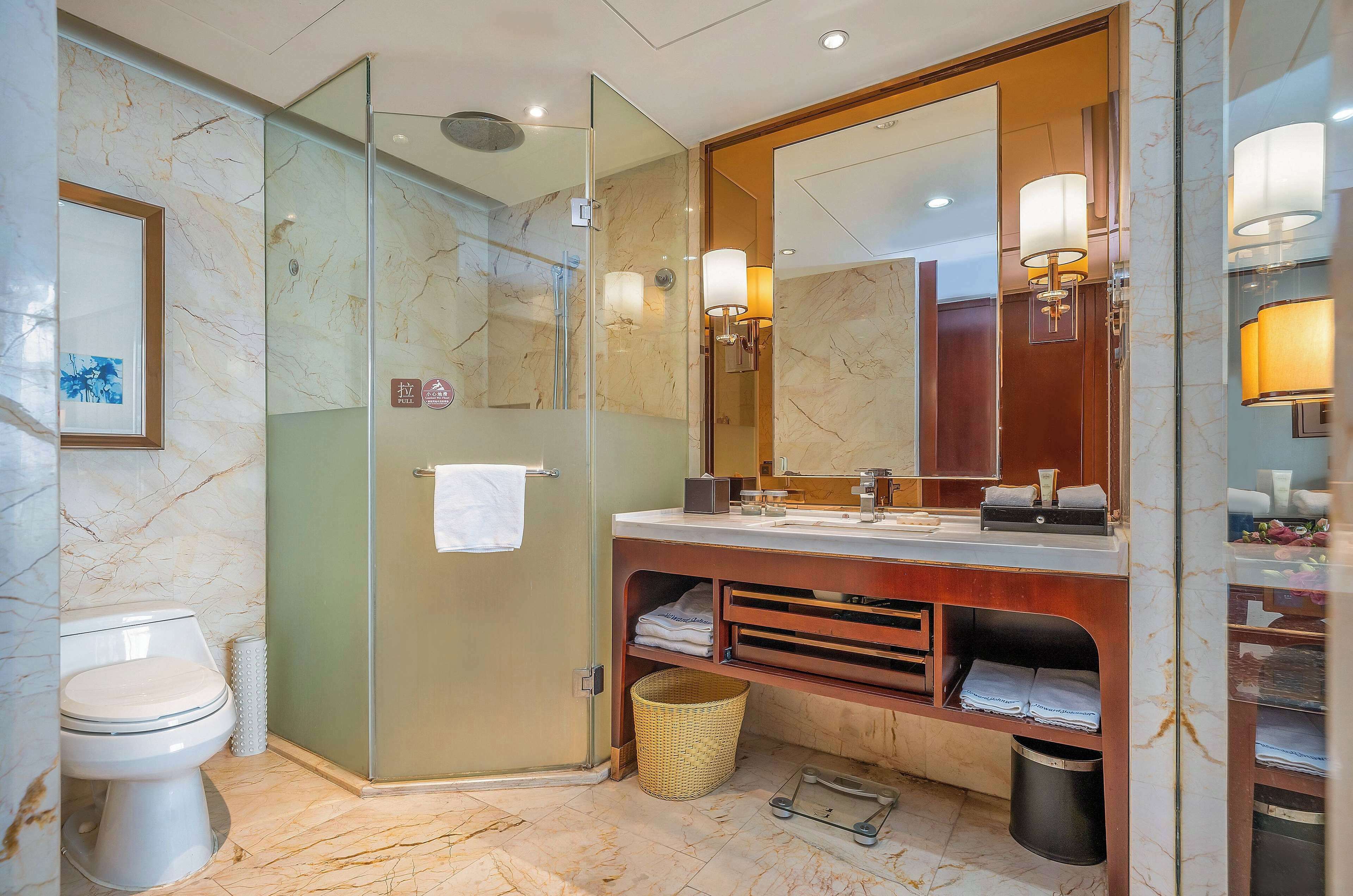 A modern bathroom with a glass-enclosed shower, a marble-tiled floor, a large mirror, and a wooden vanity with a sink and towel rack.