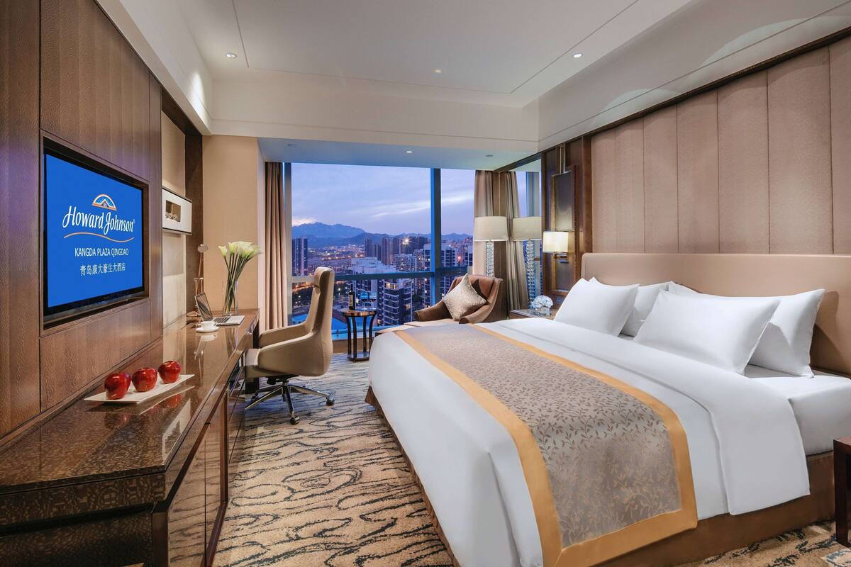 Habitación de hotel con una cama grande, un escritorio con una computadora portátil, un televisor en la pared y vista a la ciudad.