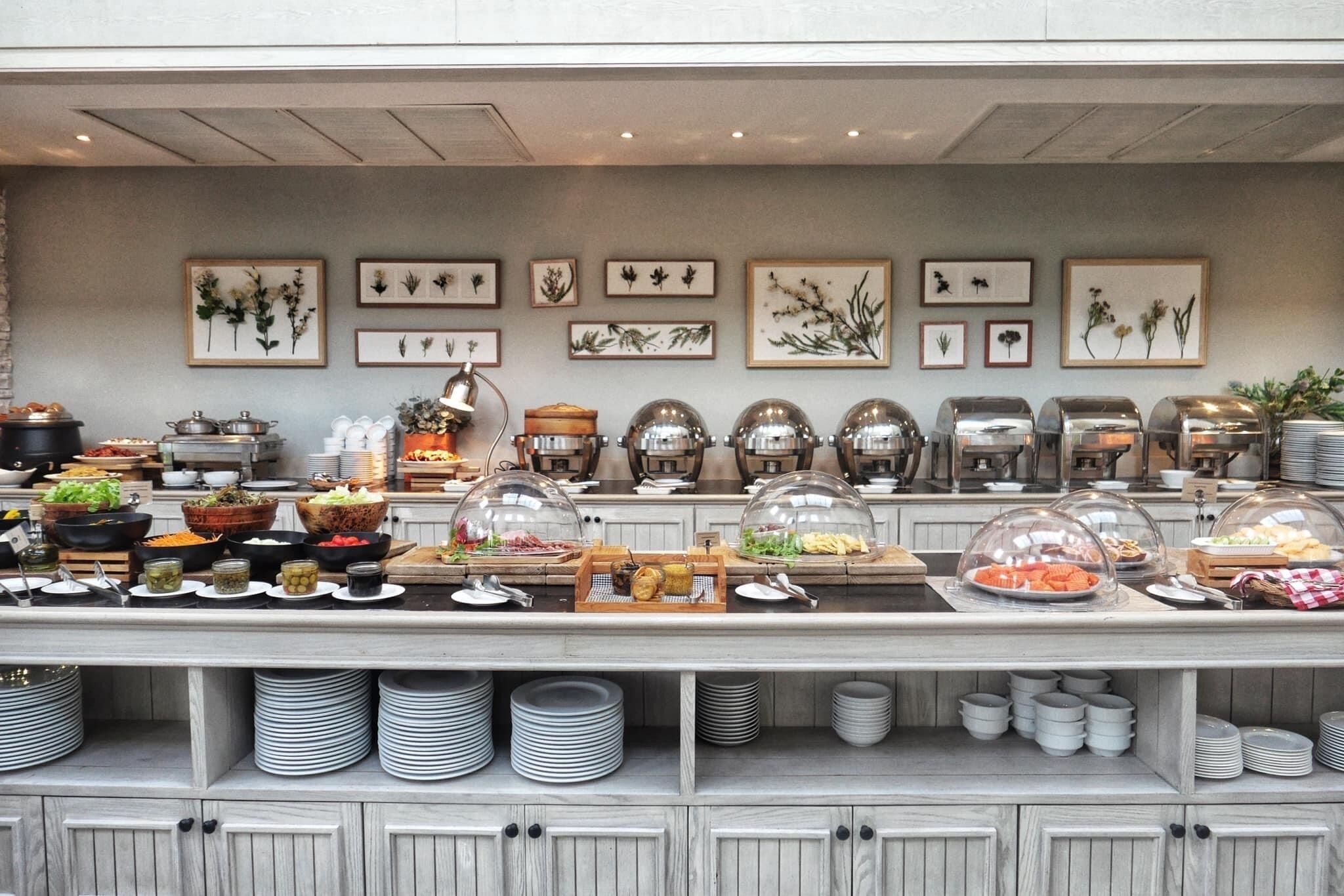 A buffet setup with various food items, serving dishes, and a wall decorated with botanical prints.