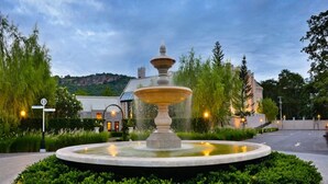 Une fontaine circulaire à plusieurs niveaux, située dans un espace paysager agrémenté d’arbustes et d’arbres.