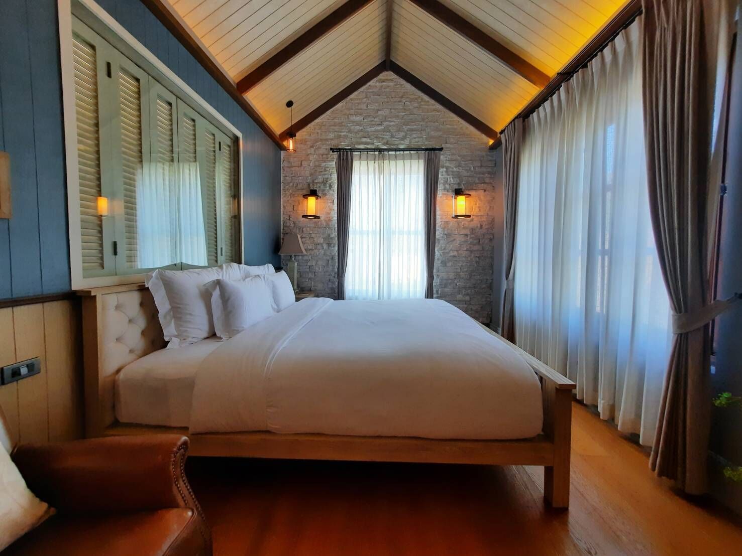 A bedroom with a large bed, wooden headboard, stone wall, and two wall-mounted lights.