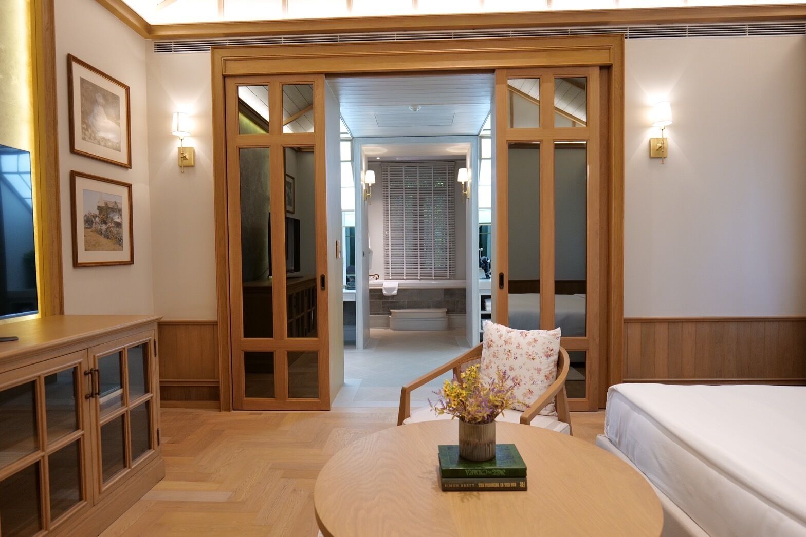 Een hotelkamer met een houten kast, een ronde tafel met een boek en bloemen, een bed, en een schuifdeur naar een badkamer.
