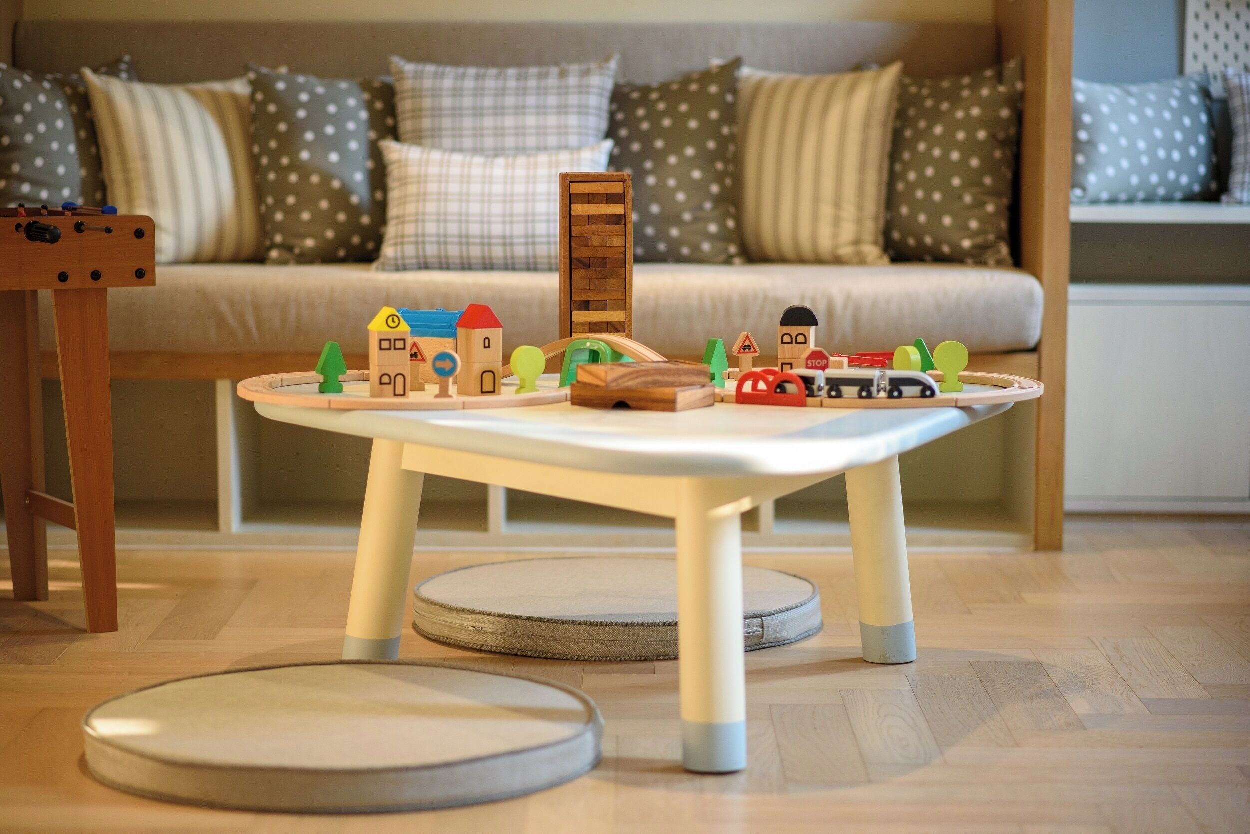 Zona infantil con una mesa, juguetes, un sofá con cojines estampados y una mesita de madera.