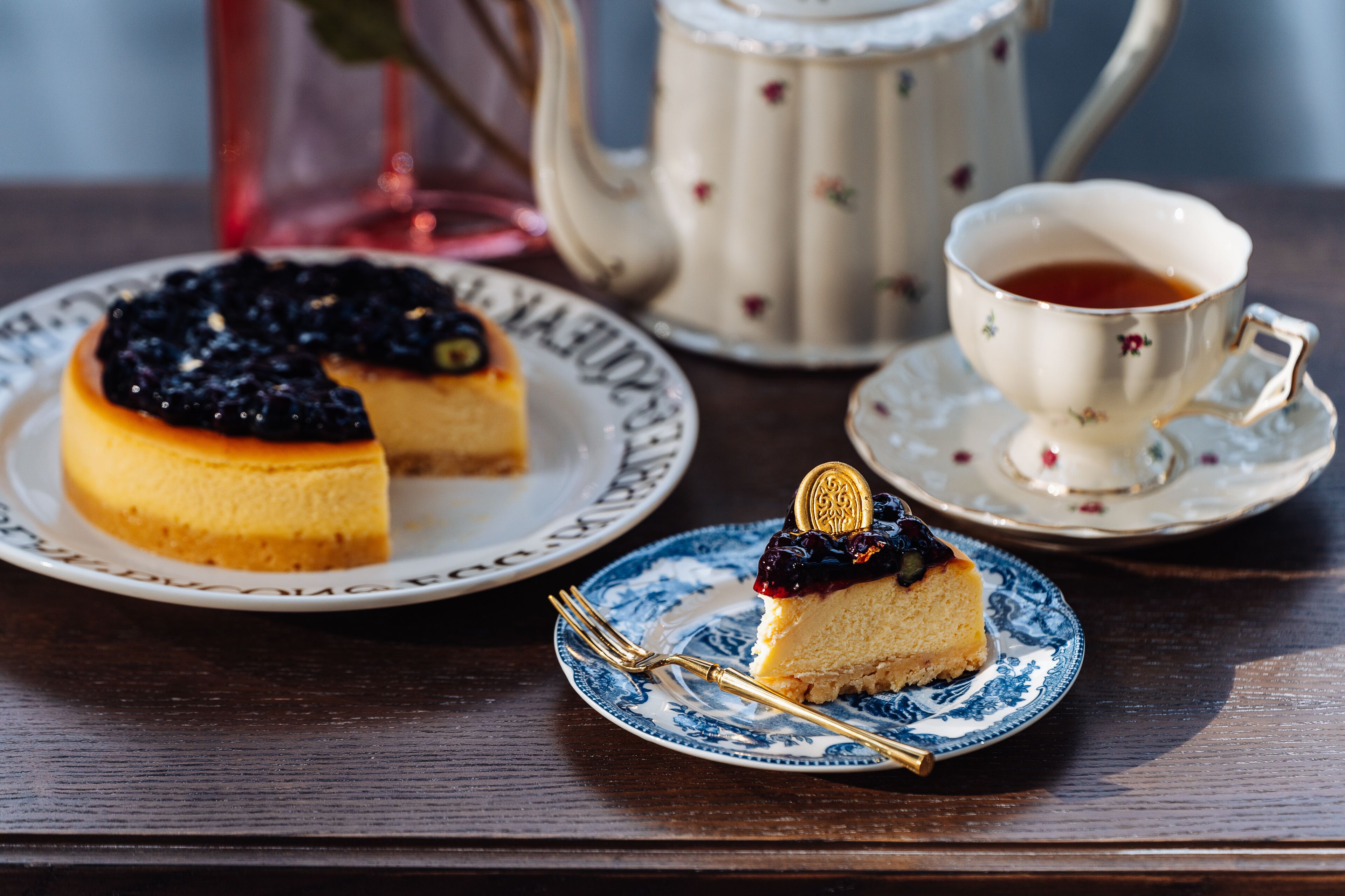 Käsekuchen auf einem Teller, eine Teetasse und ein Teekessel auf einem Holztisch.