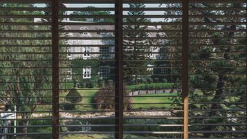 A view through blinds of a house with a well-maintained lawn, a pond, and a stone building in the background.