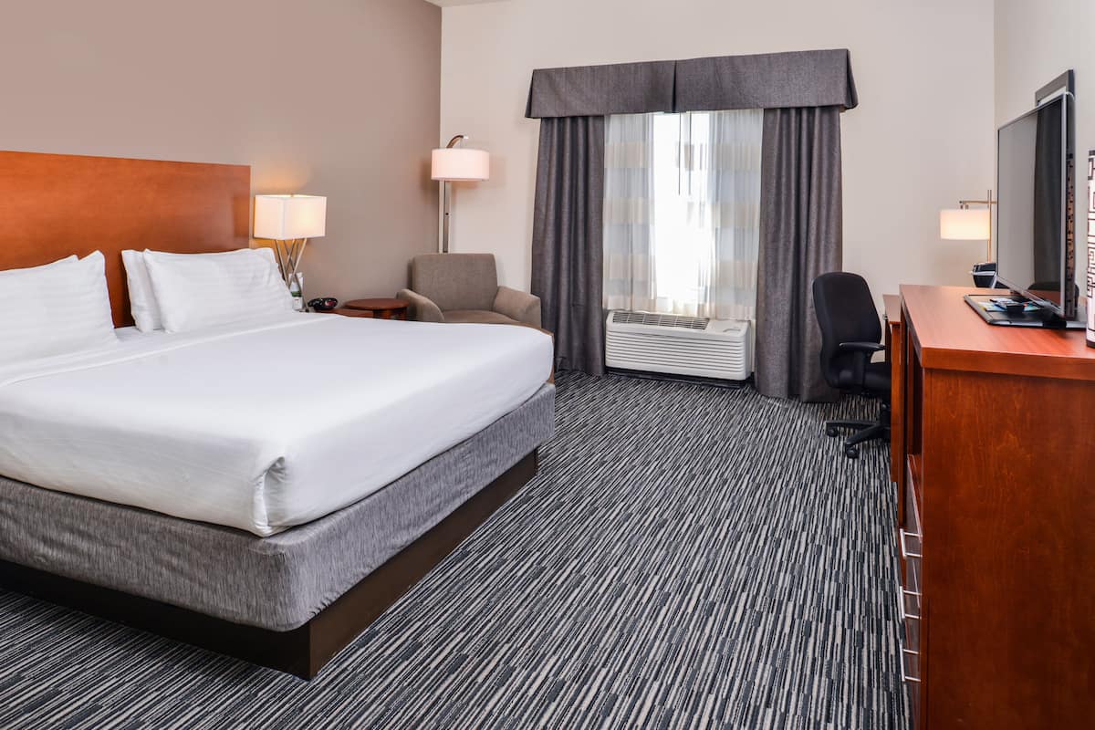A hotel room with a large bed, a desk, a chair, a lamp, and a large window with curtains.