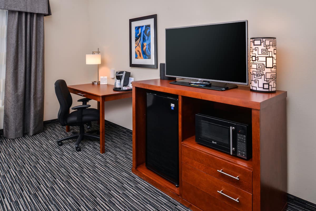 A hotel room with a wooden desk, a black refrigerator, a microwave, a black office chair, a desk lamp, and a framed abstract painting on the wall.
