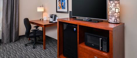 A hotel room with a wooden desk, a black refrigerator, a microwave, a black office chair, a desk lamp, and a framed abstract painting on the wall.