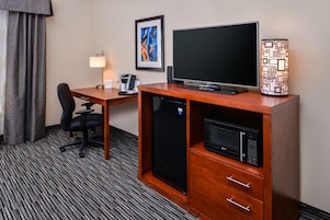 Quarto de hotel com secretária de madeira, frigorífico preto, micro-ondas, cadeira de escritório preta, candeeiro de mesa e quadro abstrato emoldurado na parede.