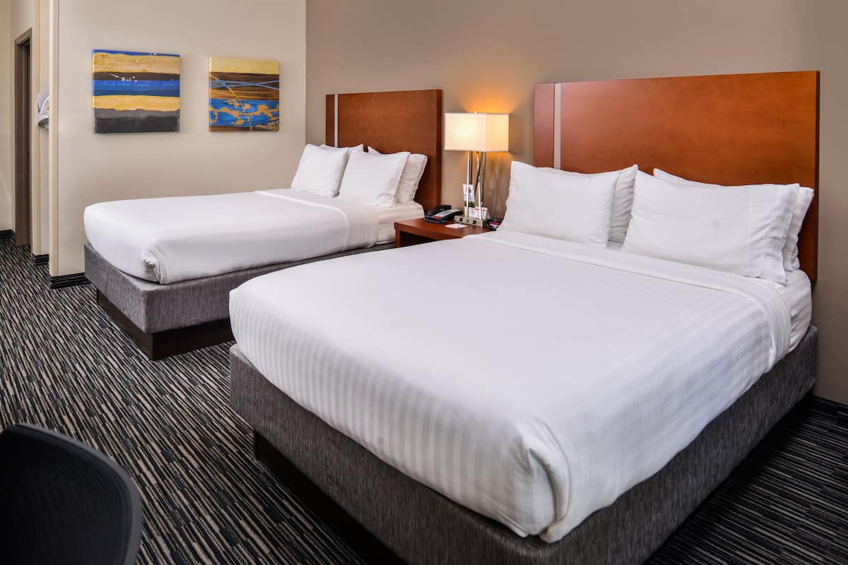 A hotel room with two beds, a nightstand with a lamp, and two paintings on the wall.