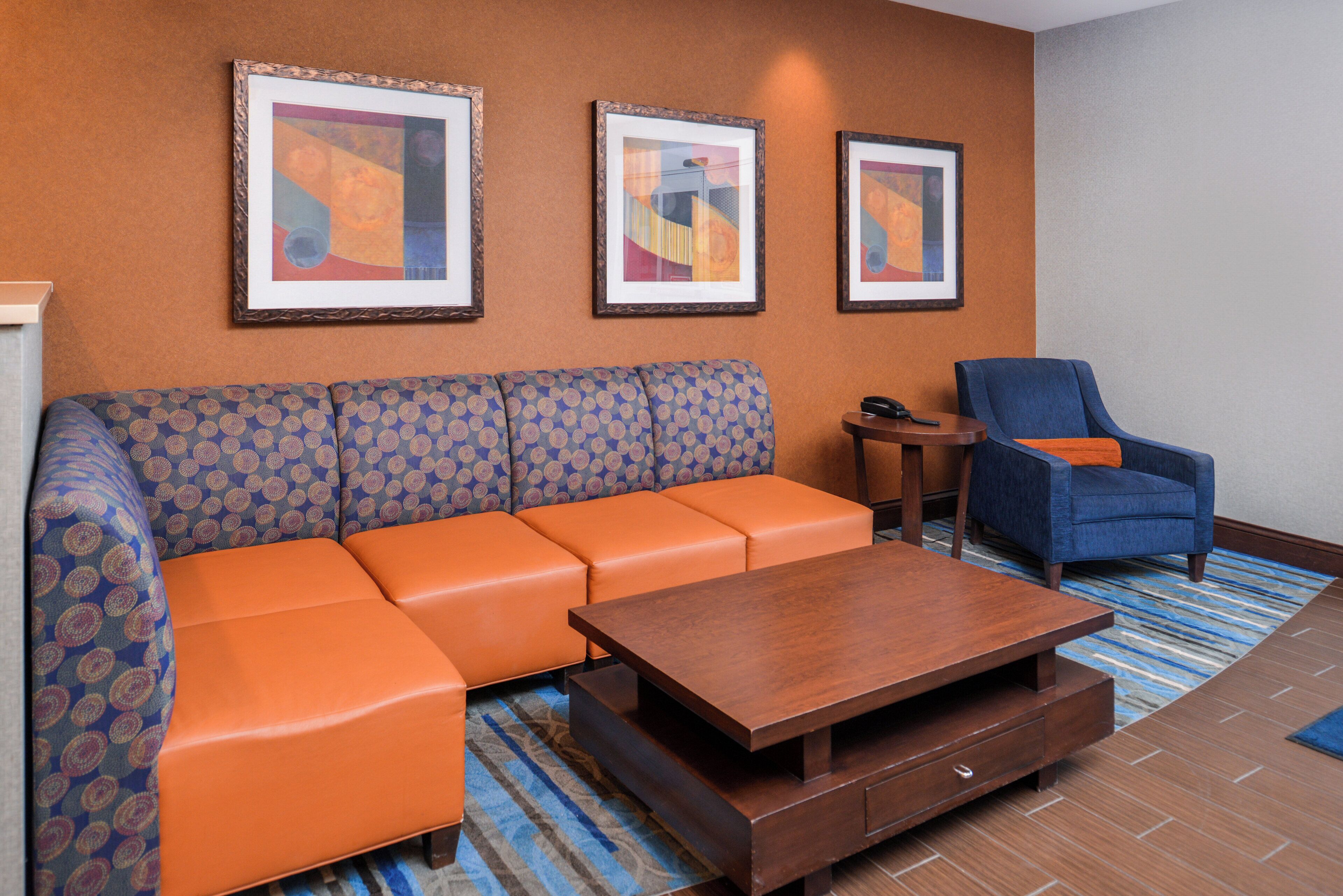 A hotel lobby with a patterned sofa, a wooden coffee table, a blue armchair, and three framed abstract paintings on the wall.