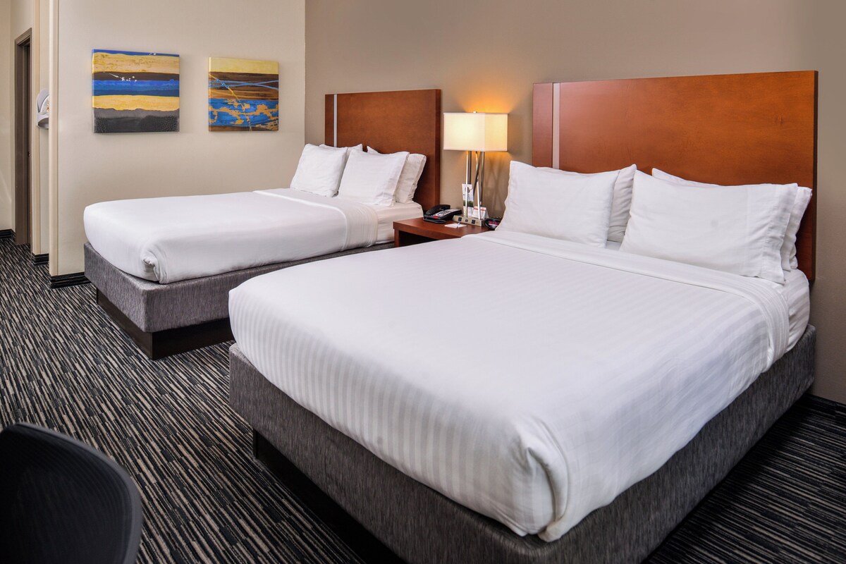 A hotel room with two beds, a nightstand with a lamp, and two paintings on the wall.