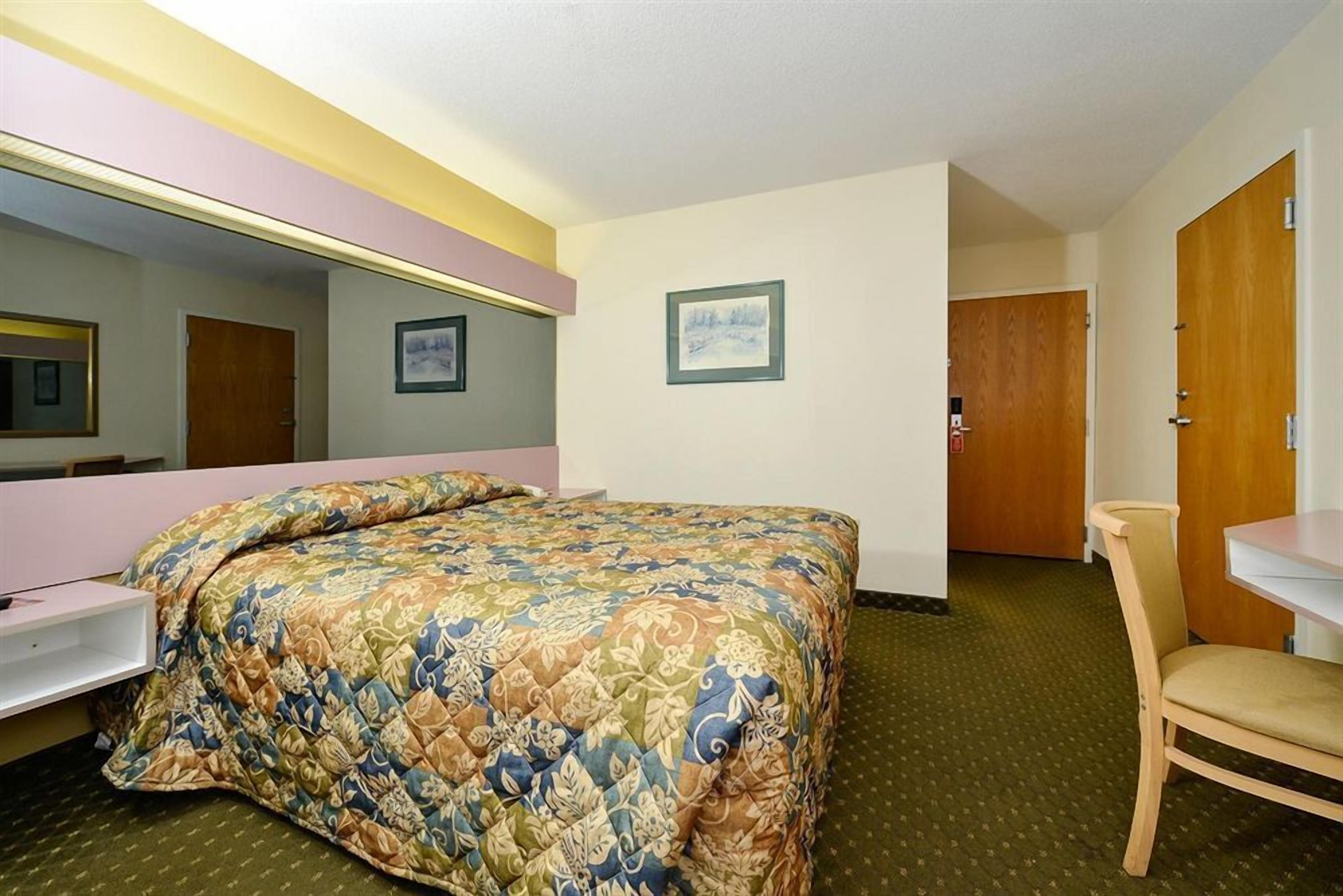 A hotel room with a bed, a chair, a desk, a mirror, and a door.