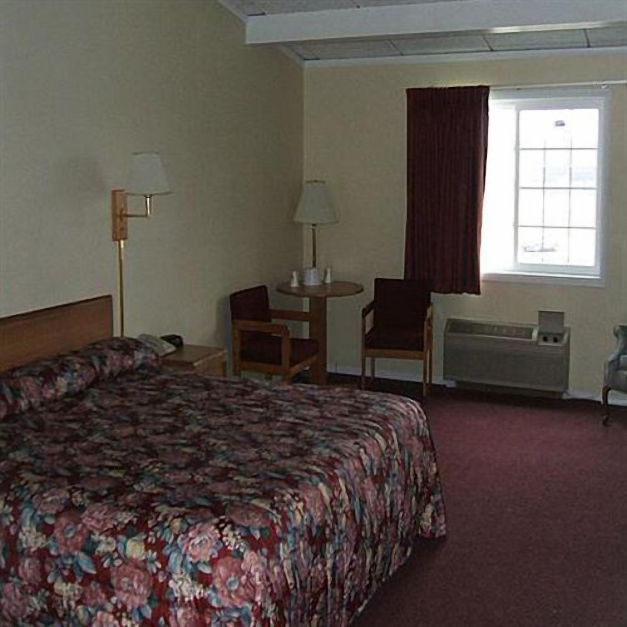 A hotel room with a bed, two chairs, a small table, a lamp, a window with curtains, and a radiator.