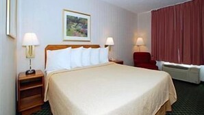 A hotel room with a bed, two bedside tables with lamps, a red chair, a window with curtains, and a framed picture on the wall.