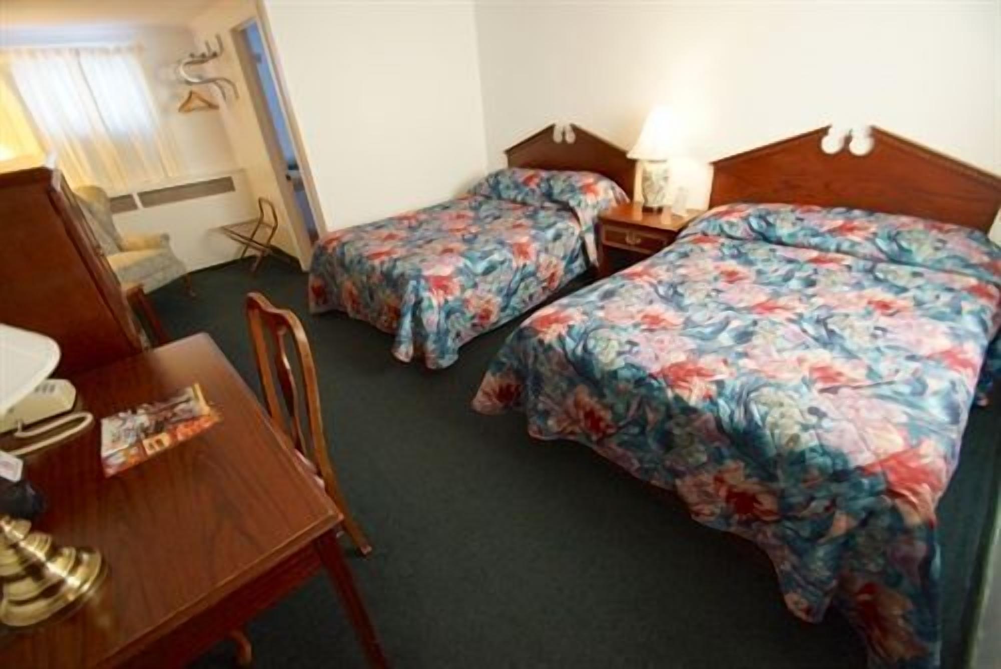 A hotel room with two single beds, a wooden desk, a chair, and a small table with a lamp.