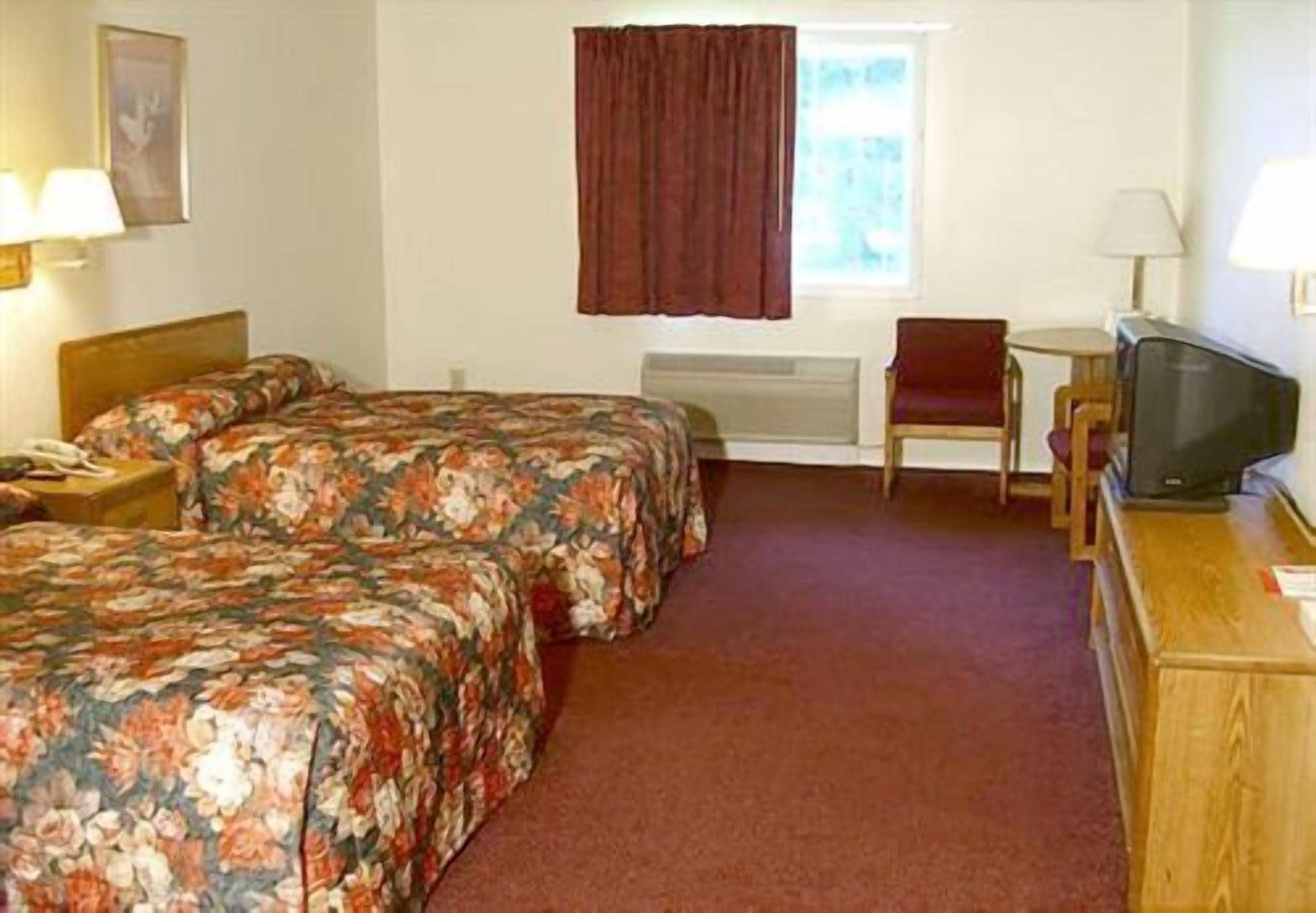 A hotel room with two beds, a small table, a chair, a TV, and a window with curtains.