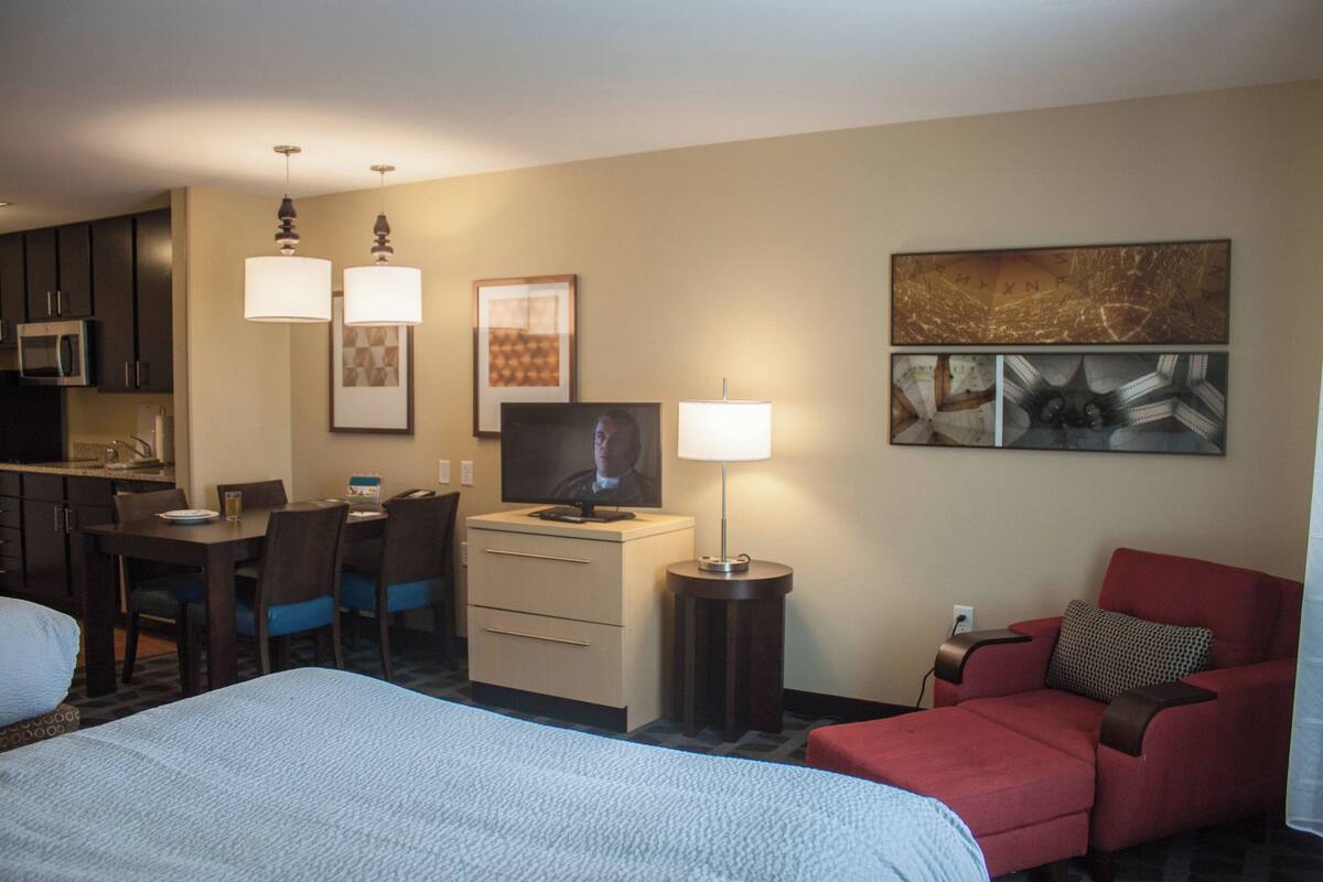Ein Hotelzimmer mit einem Bett, einem Schreibtisch mit Computer, einem Fernseher, einer Lampe und einem roten Sessel.