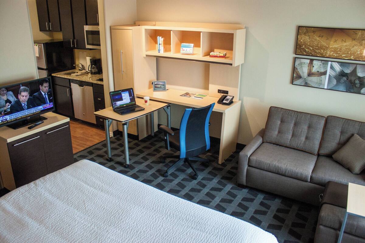 Ein Hotelzimmer mit Bett, Schreibtisch mit Laptop, Sessel, Sofa, Mikrowelle, Kühlschrank und Fernseher, der eine Nachrichtensendung zeigt.
