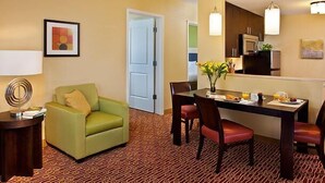 Habitación de hotel con comedor, un sillón verde, una mesa con sillas, una lámpara y obras de arte en las paredes.