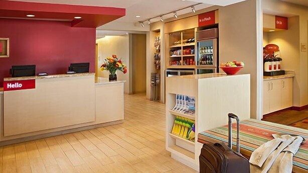 Zona de recepción de hotel con mostrador, un letrero que dice "Hello", un pequeño área de descanso con mesa y sillas, y una kitchenette con cafetera.