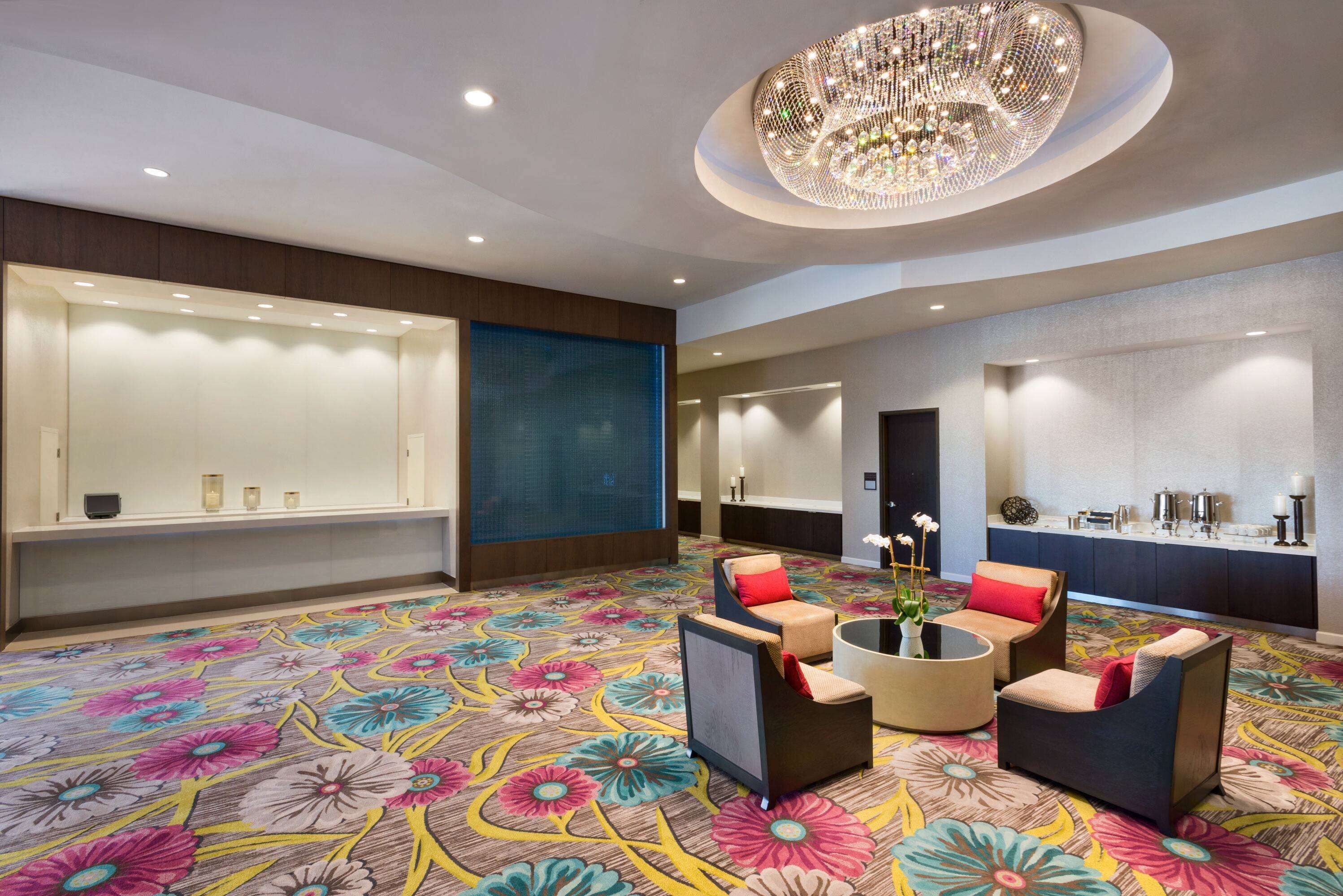 A hotel lobby with a floral carpet, seating area with armchairs and a coffee table, a reception desk, and a decorative chandelier.