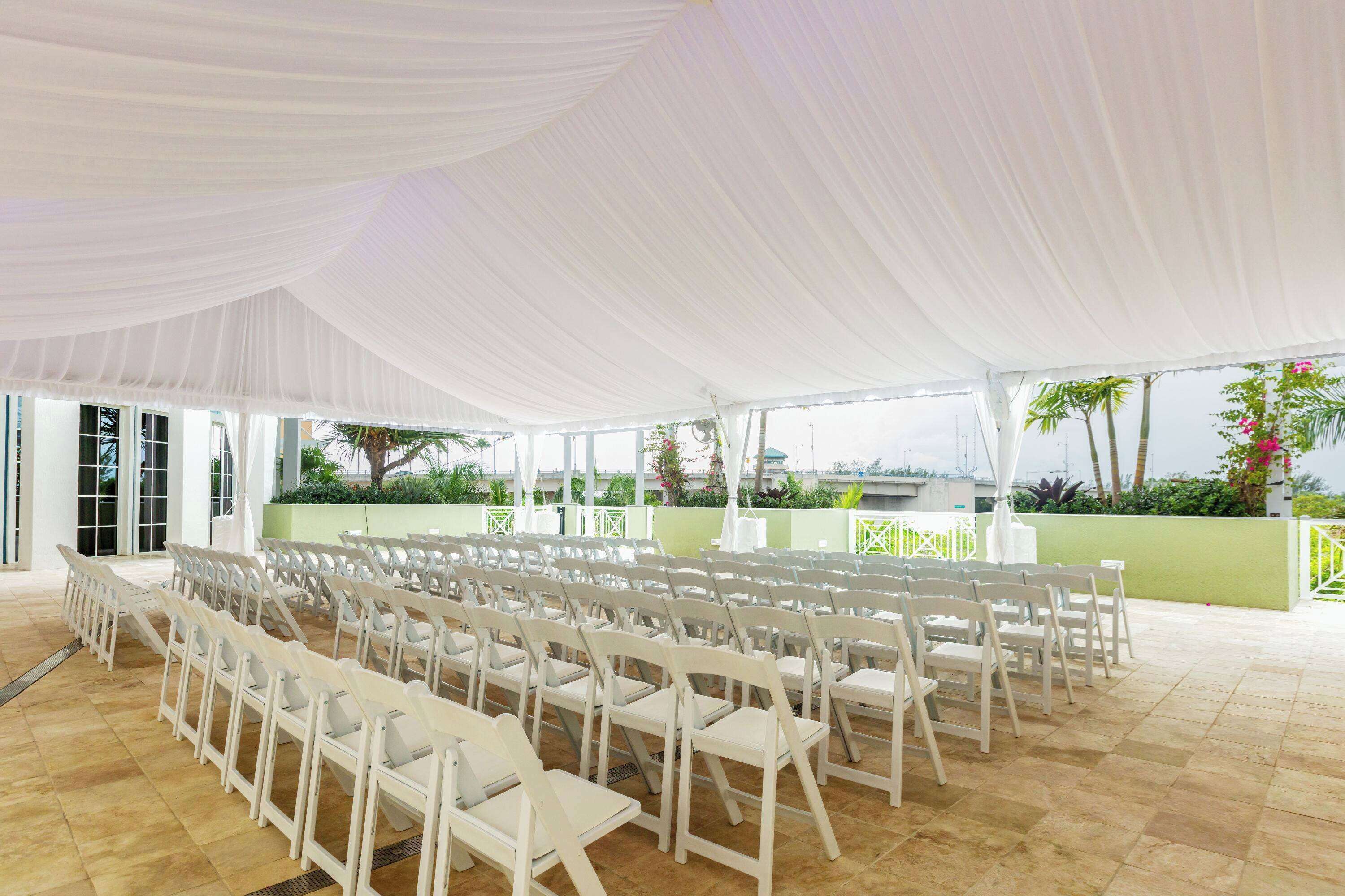 An indoor event space with rows of white chairs arranged for seating, large white drapes covering the ceiling, and a view of greenery and buildings outside.