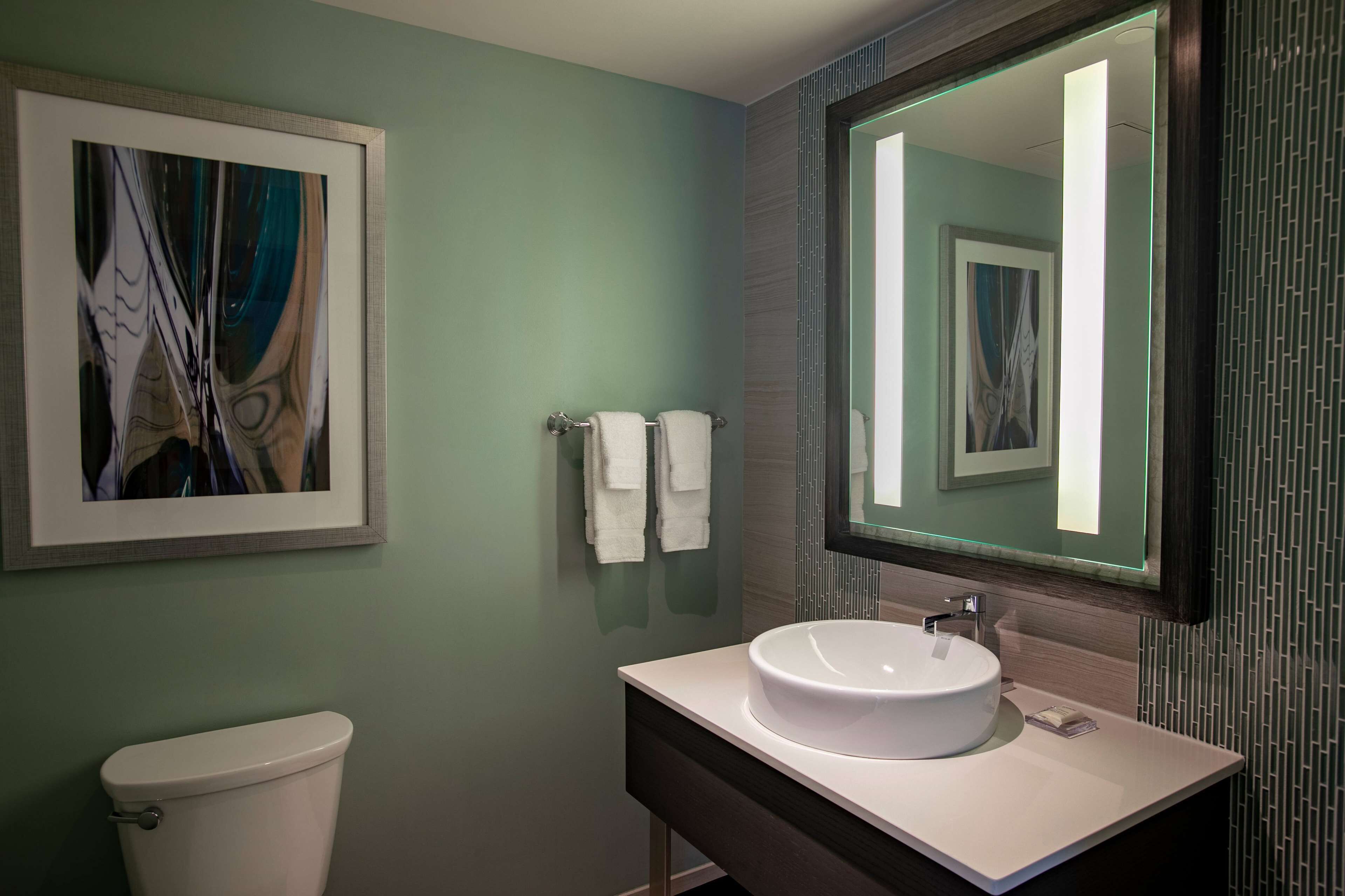 A modern bathroom with a white toilet, a large framed abstract painting, a round sink with a mirror above it, and two white towels hanging on a towel rack.
