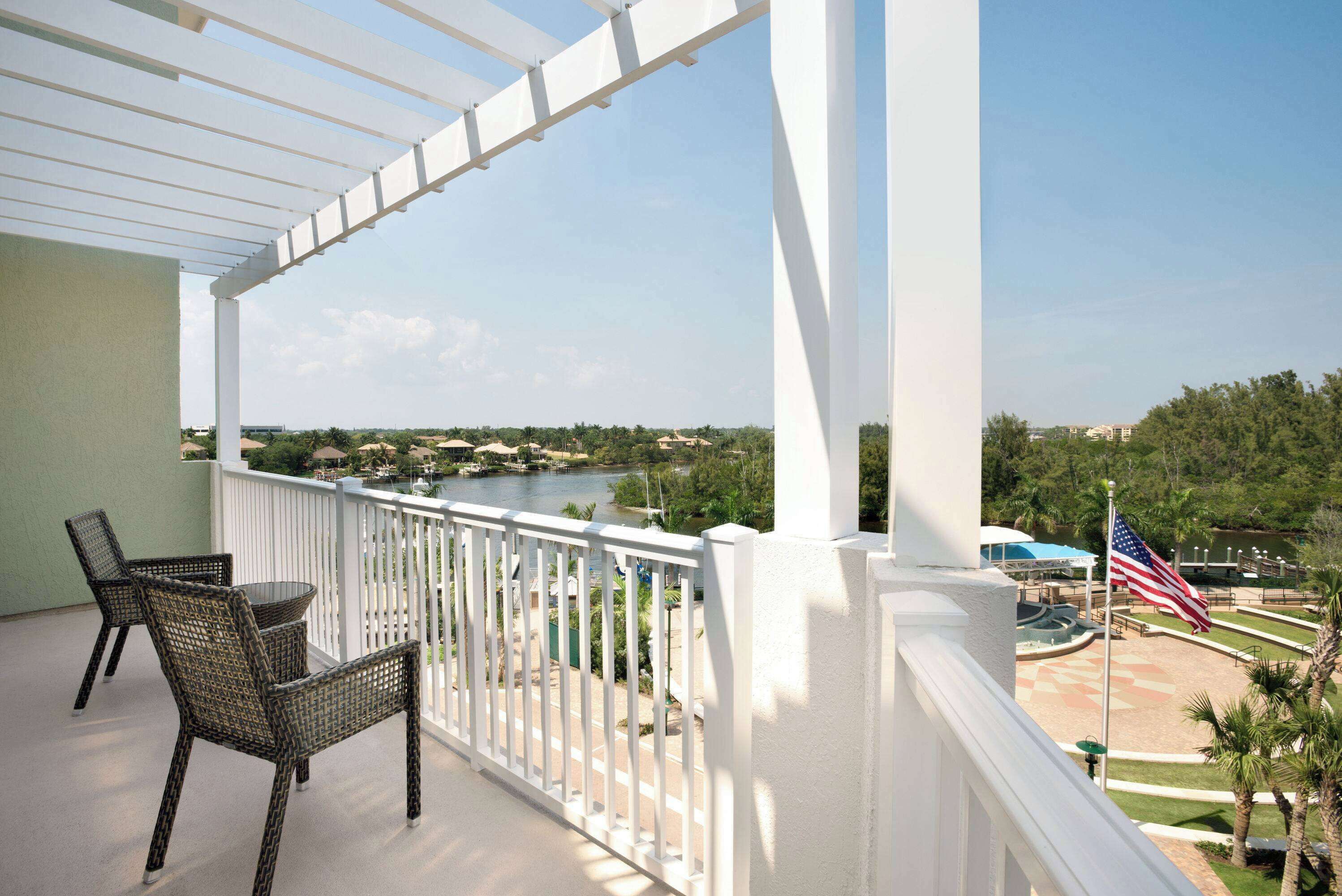 A balcony with wicker chairs and a table, overlooking a river and a park with an American flag.