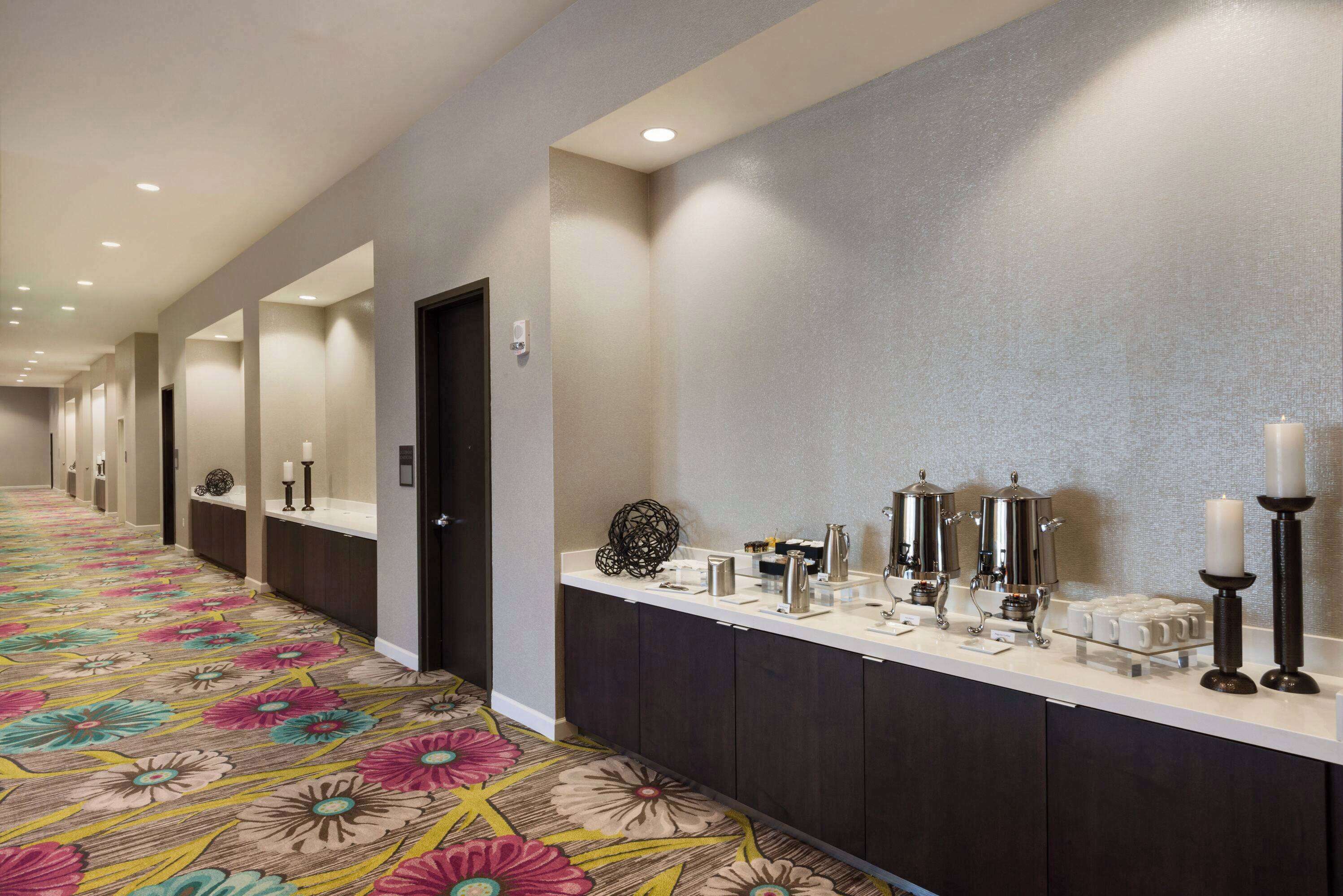 A hotel corridor with a floral carpet, a long reception desk with coffee machines, and a mirror wall.