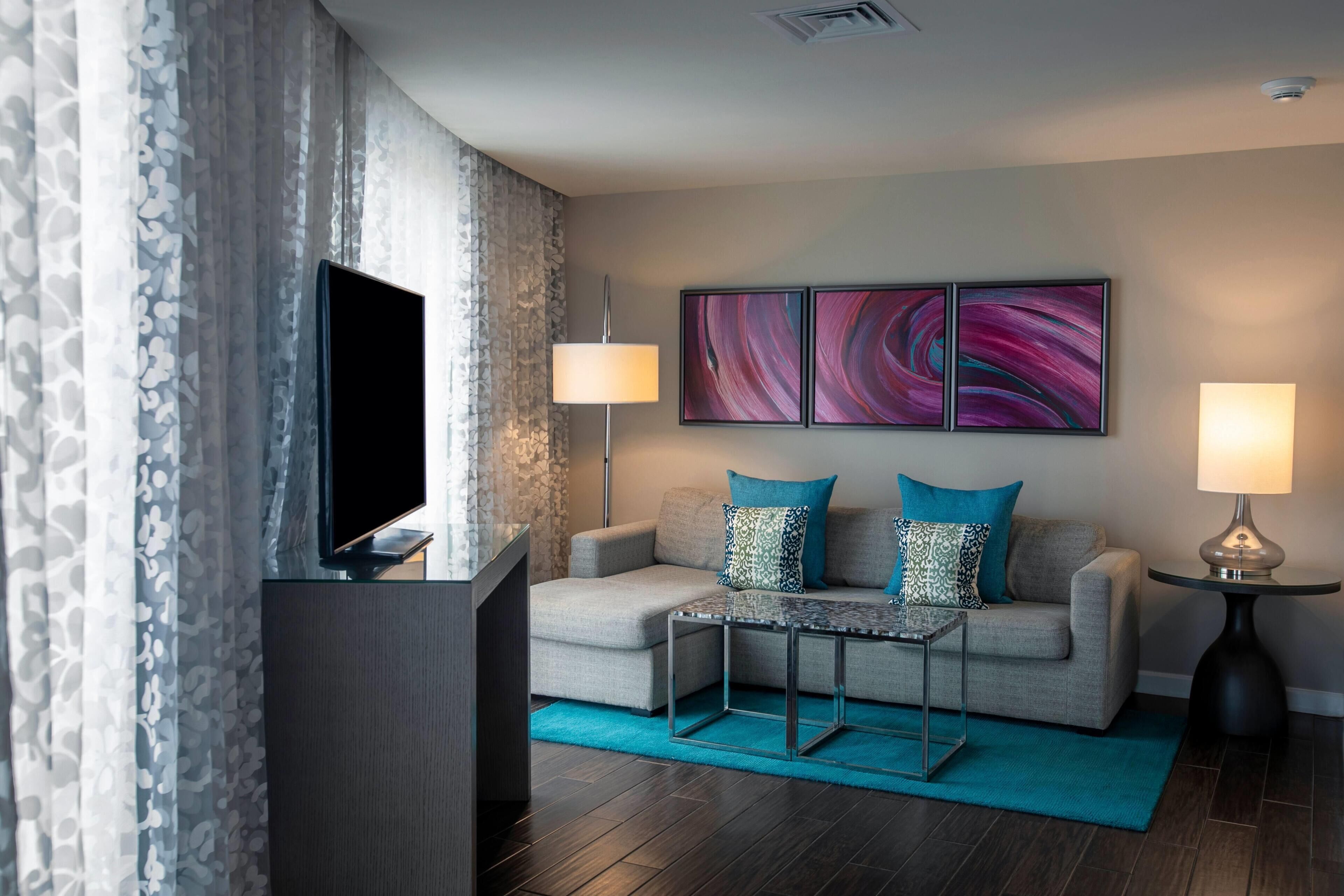 A modern living room with a flat-screen TV, a grey sofa with patterned cushions, a coffee table, and two floor lamps.