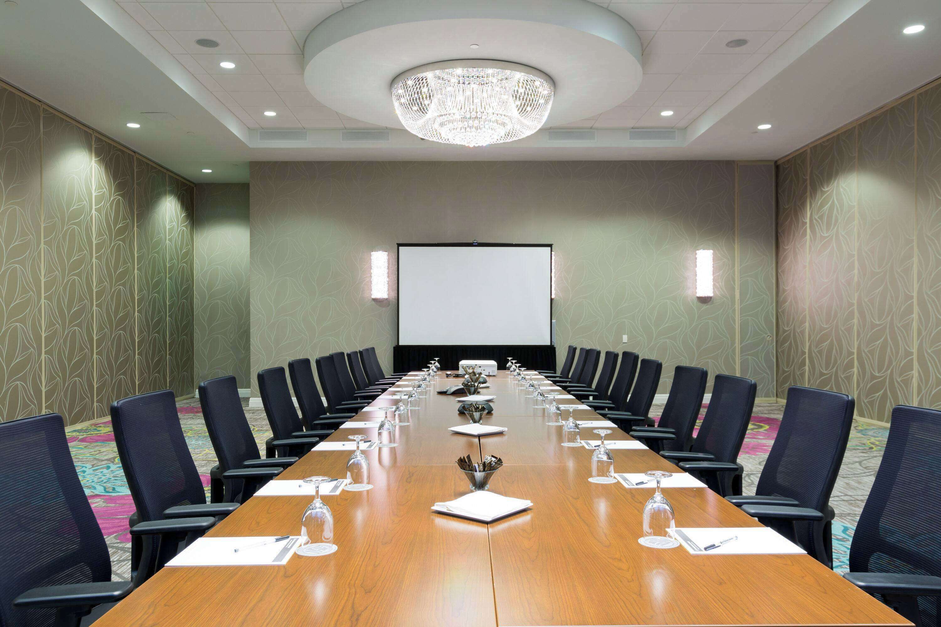 A conference room with a long table, chairs, a projector screen, and a chandelier.