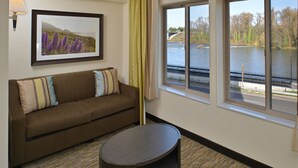 A hotel room with a brown sofa, a small round table, a framed landscape picture on the wall, and a window offering a view of a river and bridge. - Candlewood Suites Eugene Springfield by IHG (Eugene)