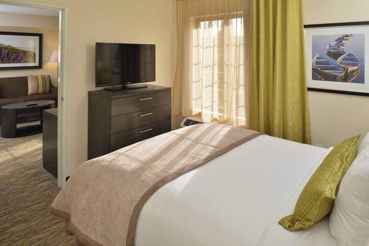 Habitación de hotel con una cama, un televisor en una cómoda, una ventana con cortinas y un cuadro en la pared.