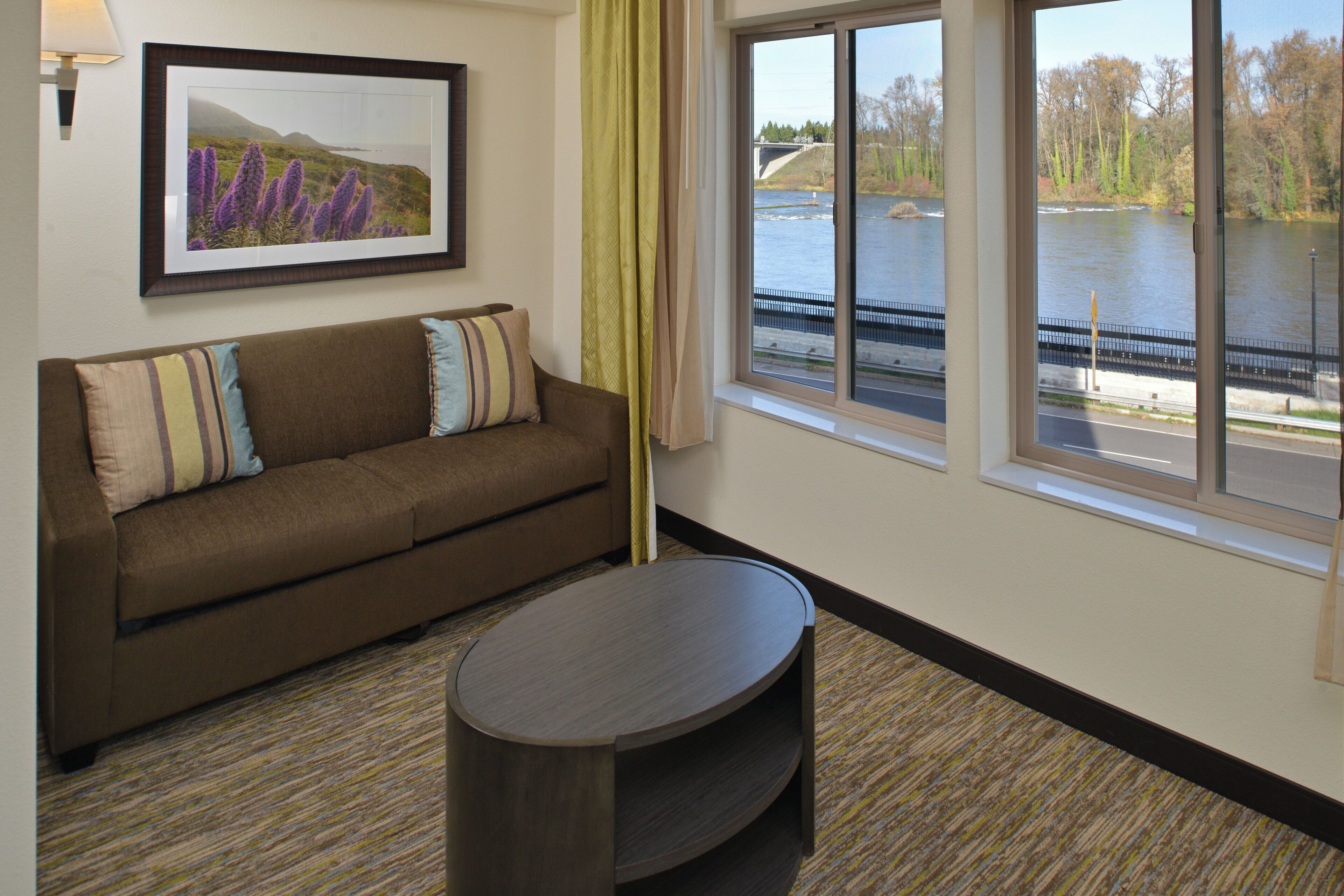 A room with a sofa, a small table, a framed picture on the wall, and a window with a view of a river and bridge.