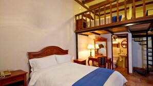 Hotelkamer met een houten bed, nachtkastje, bureau met lamp, spiegel en een zichtbare badkamer door een open deur.